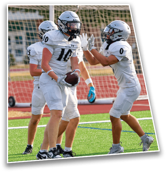 Three people in white football uniforms and helmets celebrate on a green field near a goalpost, with one holding a football and another reaching out for a high-five. Description generated by AI