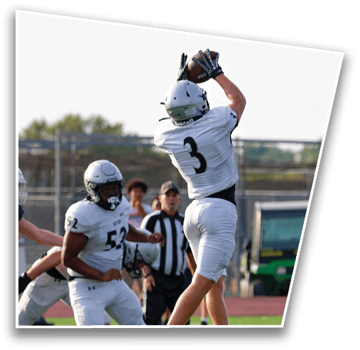 A person wearing a white football uniform with the number 3 is jumping to catch a football while another person in a white uniform with the number 52 watches nearby on a football field. Description generated by AI