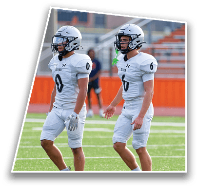 Two people wearing white football helmets and white jerseys stand side by side on a sports field with a blurred background of buildings and bleachers. Description generated by AI