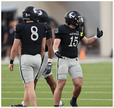 Three people in football uniforms stand on a green field with one person gesturing while wearing a helmet and gloves. Description generated by AI