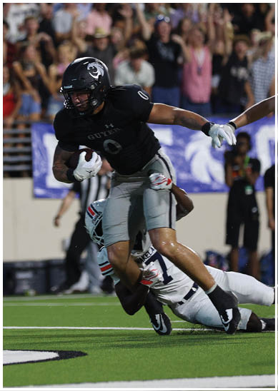 A person in a black football uniform is running with the ball while being tackled by a person in a white uniform on a football field with a crowd in the background. Description generated by AI