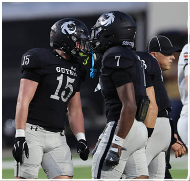 Two football players in black uniforms with helmets are facing each other closely on the field while a coach or official stands nearby. Description generated by AI
