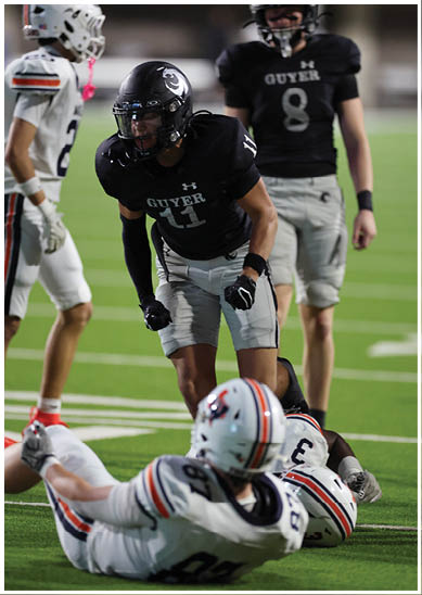 An adult wearing a black football uniform with the word “GUYER" is standing on a field while two adults in white football uniforms with helmets are lying on the ground. Description generated by AI