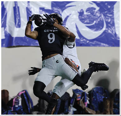 Two people in football uniforms are engaged in a play with one person attempting to catch the ball while the other tries to block or tackle. Description generated by AI