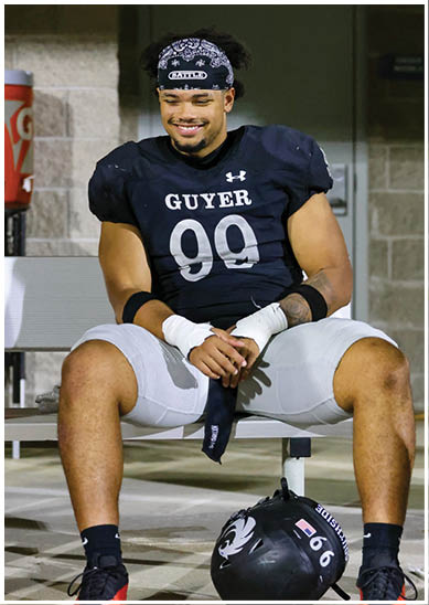 A person wearing a black football jersey with the number 99 and the word Guyer is sitting on a bench with a black helmet on the ground in front. Description generated by AI
