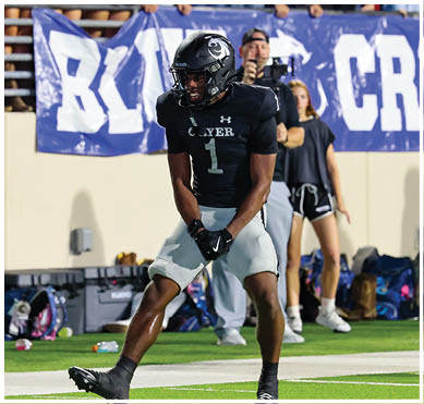 A person wearing a black football uniform with the number one is standing on a field with a banner and other people in the background. Description generated by AI