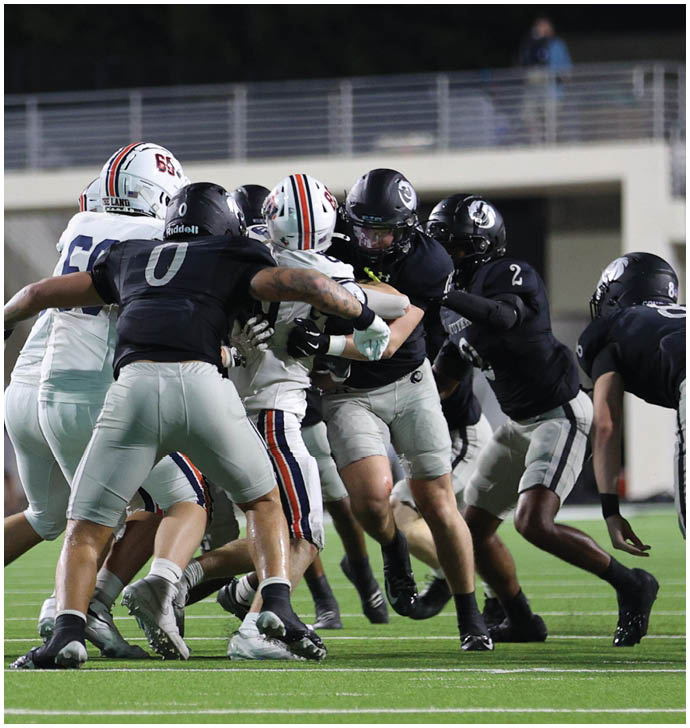 Several football players in black and white uniforms are engaged in a close tackle on a green field during a game. Description generated by AI
