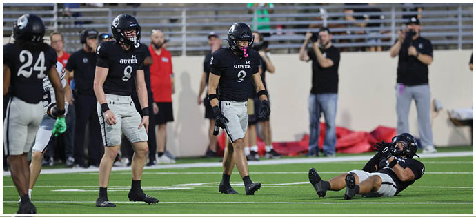 Several people in football uniforms are on a field with one person lying on the ground while others stand nearby and spectators watch from the bleachers. Description generated by AI