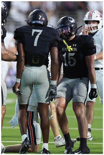 Two people wearing black football uniforms and helmets are engaged in conversation on a field with a blurred crowd in the background. Description generated by AI