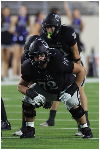 Three people wearing black football uniforms and helmets are positioned closely together on a field, appearing focused and ready for action. Description generated by AI