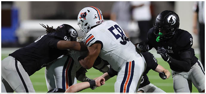 Three people in football uniforms are engaged in a tackle during a game on a field. Description generated by AI