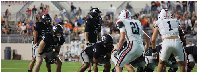 People in football uniforms are positioned on a field preparing for a play with a crowd visible in the background. Description generated by AI