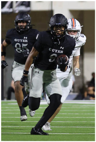 A person wearing a black football uniform with the word “GUYER" is running with a football while another person in a similar uniform and a person in a white uniform chase behind on a football field. Description generated by AI