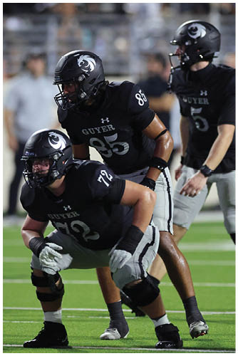 Three football players in black uniforms with helmets are positioned on a field, appearing ready for a play during a game. Description generated by AI