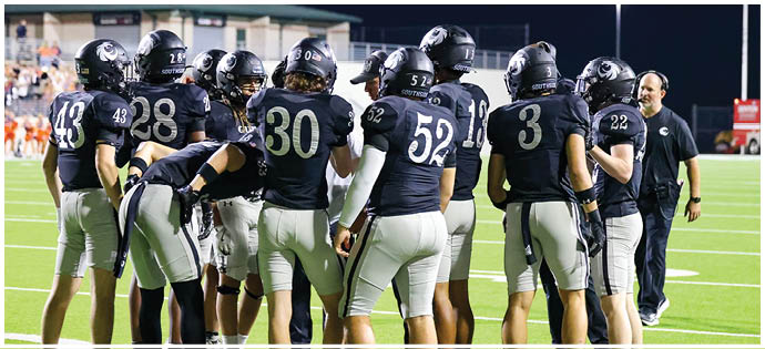 A group of football players in black jerseys and white pants are gathered on a field at night with a coach standing nearby. Description generated by AI