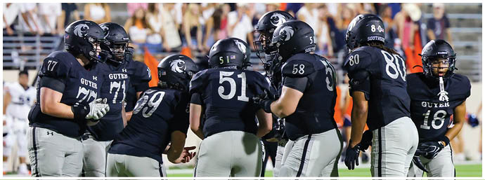 A group of people wearing black football uniforms with white pants are huddled together on a green field during a game. Description generated by AI