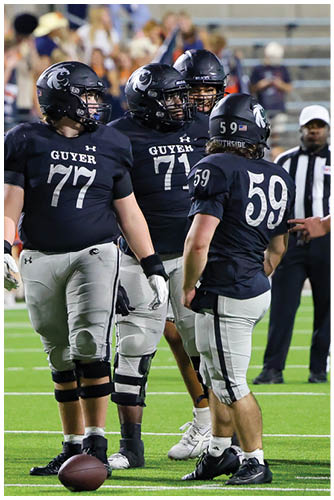 People wearing black football uniforms with helmets are standing on a green field with a referee in the background. Description generated by AI