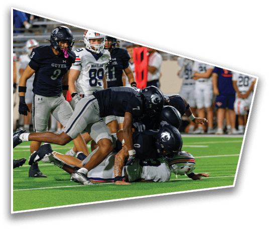 Several football players in dark uniforms are tackling a player in a white uniform on a green field during a game. Description generated by AI