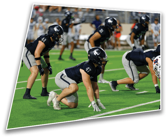 Several people in dark sports uniforms and helmets are positioned on a green football field, preparing for a play. Description generated by AI