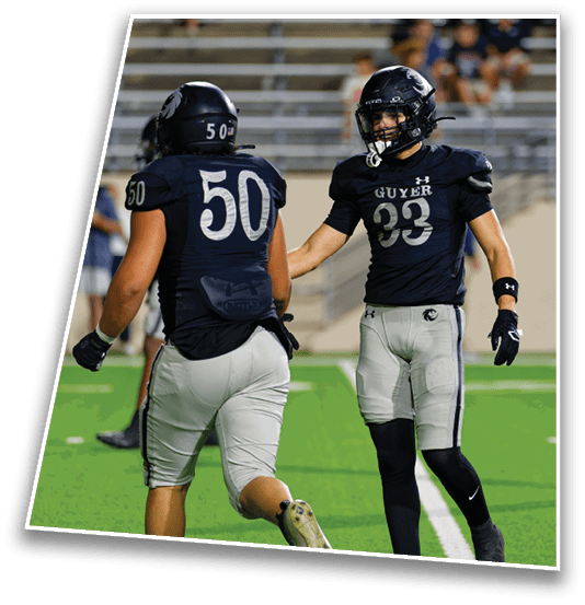 Two people wearing football uniforms with helmets are on a green field, one facing forward and the other with their back to the camera. Description generated by AI