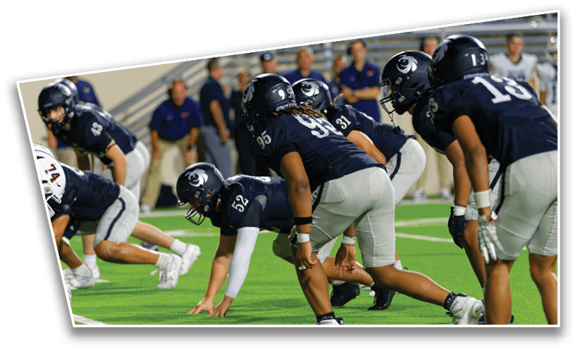 Several football players in dark jerseys and helmets are lined up on a green field preparing for a play during a game. Description generated by AI