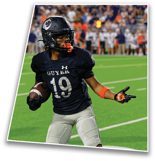 A person wearing a black football uniform with the number 19 and the word Guyer is holding a football and pointing with their right hand on a green field with blurred spectators in the background. Description generated by AI