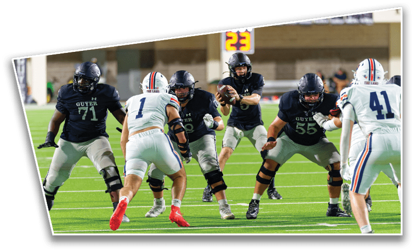 Players in football uniforms are engaged in a play on a green field with one player holding the ball behind the offensive line while others prepare to block or defend. Description generated by AI