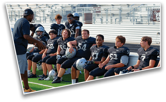 A group of people in football uniforms sit on a bench on a sports field while a coach stands nearby giving instructions. Description generated by AI