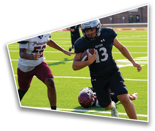 A person wearing a dark sports uniform is running on a football field while holding a football, with another person in a white and maroon uniform nearby and a helmet on the ground. Description generated by AI
