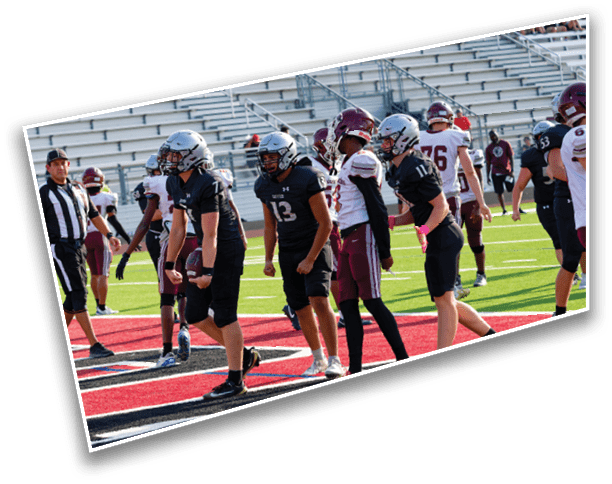 Several people wearing football uniforms and helmets stand on a field near the end zone with empty bleachers in the background. Description generated by AI