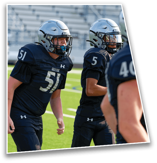 Three people wearing black football uniforms and silver helmets stand on a green field with bleachers in the background. Description generated by AI