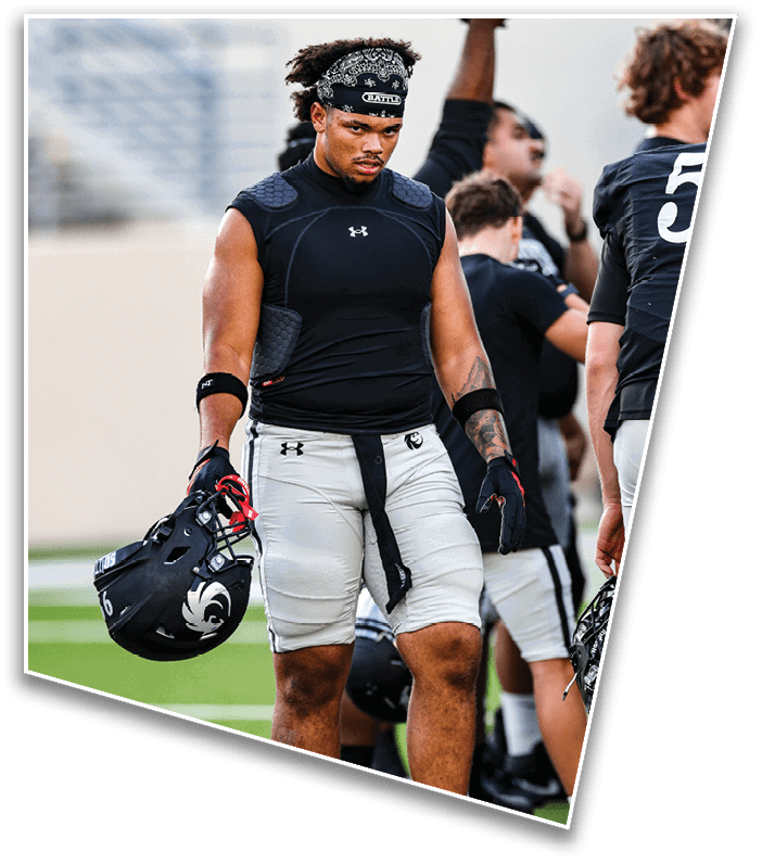 A person wearing a black sleeveless compression shirt and white football pants is holding a black football helmet and standing on a field with other people in sports attire in the background. Description generated by AI