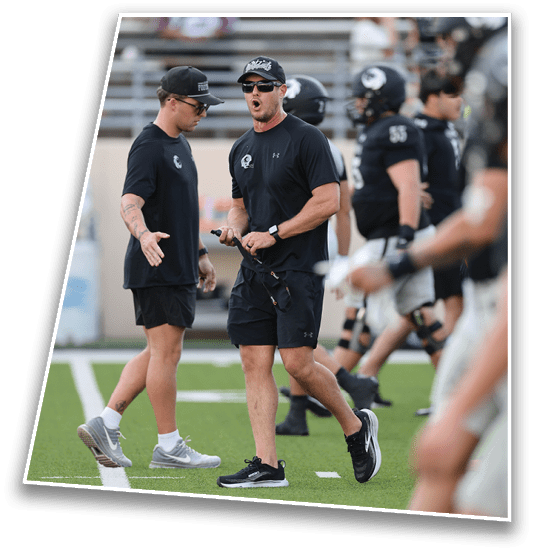 Two adults wearing black athletic clothing and caps are engaged in a discussion on a football field with players in the background. Description generated by AI