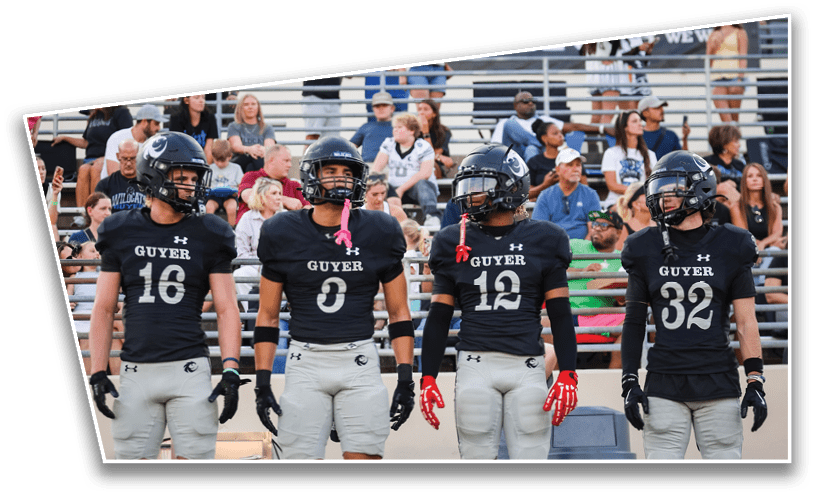 Four people wearing black football jerseys and helmets stand side by side on a field with spectators in the background. Description generated by AI