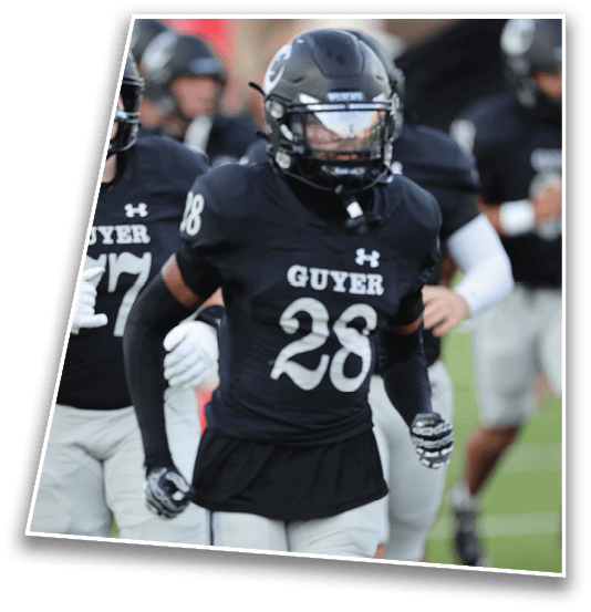 A person wearing a black football uniform with the number 28 and the word “GUYER" on the front is running on a field with other players in similar uniforms in the background. Description generated by AI