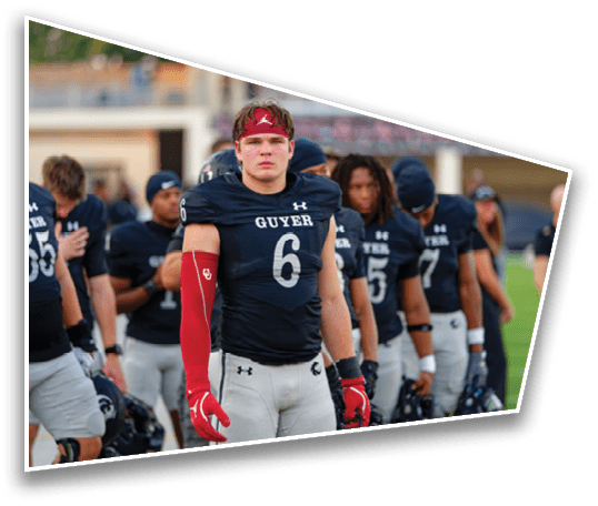 A group of athletes in football uniforms stand in a line on a field with one athlete in the foreground wearing a red arm sleeve and headband. Description generated by AI