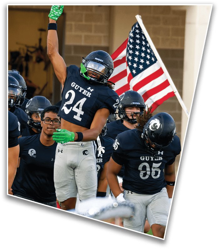 Several people in football uniforms and helmets are on a field with one person holding an American flag in the background. Description generated by AI