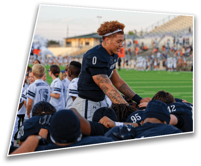 A person in a dark sports uniform with the number zero is standing and interacting with teammates who are kneeling on a football field with a stadium and spectators in the background. Description generated by AI