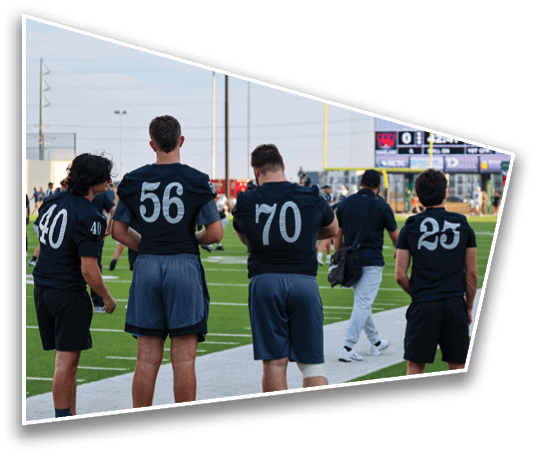 Four people wearing black sports jerseys with numbers stand on the sidelines of a football field while another person walks nearby. Description generated by AI