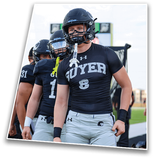 People wearing black football helmets and black athletic shirts stand together outdoors with a scoreboard visible in the background. Description generated by AI