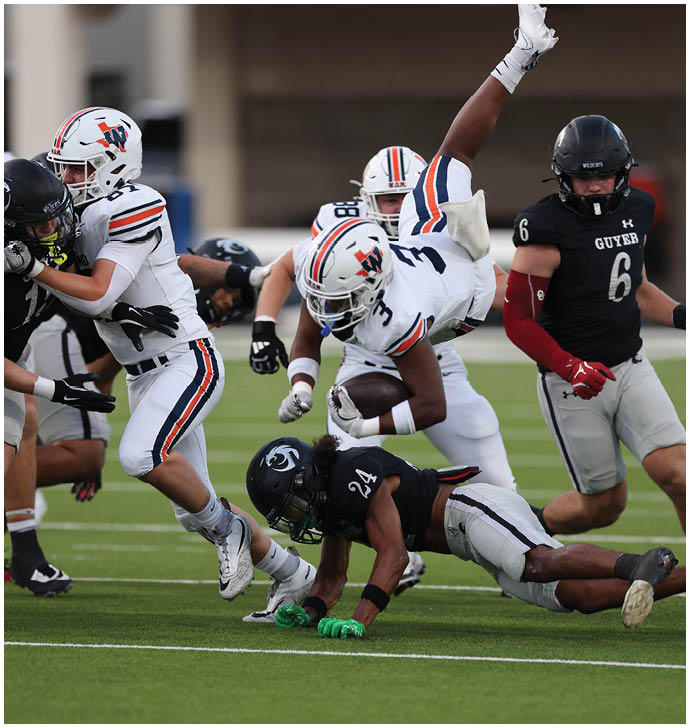 A group of people in football uniforms are actively engaged in a play on a green field with one person in white uniform holding the ball while others in black and white uniforms attempt to tackle or block. Description generated by AI