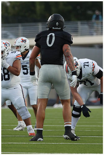 Four people in football uniforms are positioned on a field with one person in a black jersey facing away and three others in white jerseys preparing for a play. Description generated by AI