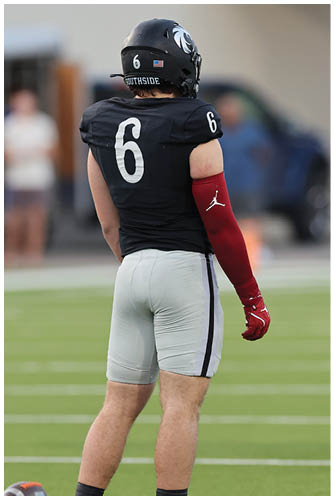 A person wearing a black football uniform with the number six on the jersey and helmet stands on a field with a red arm sleeve featuring a white logo. Description generated by AI