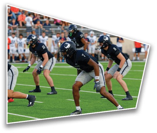 Several people in dark sports uniforms and helmets are positioned on a football field, appearing ready for a play during a game. Description generated by AI