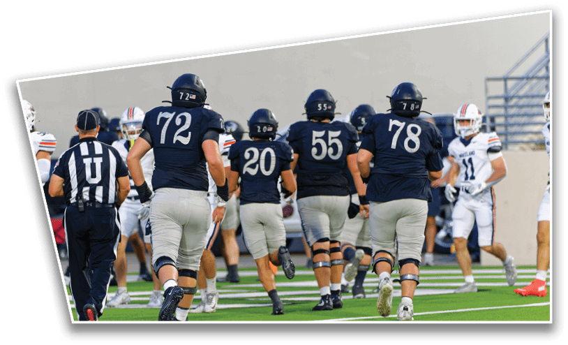 Several people in dark sports uniforms and helmets are running on a football field toward others in white uniforms while a referee walks nearby. Description generated by AI