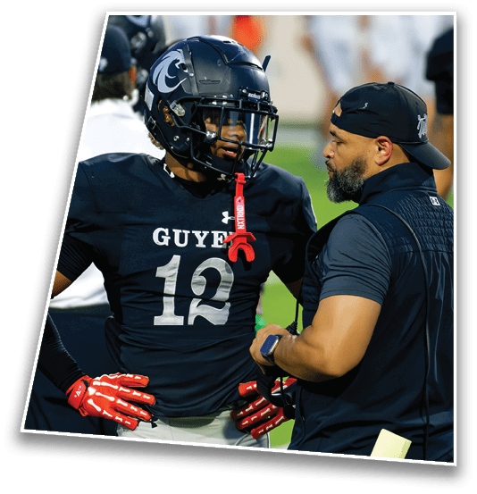 A person wearing a football uniform with the number 12 and the word “GUYER" on the jersey is standing with hands on hips while talking to another person dressed in black sportswear on a football field. Description generated by AI