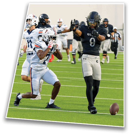 Two people in football uniforms are actively engaged on a field with one person in a black uniform signaling with their hands and another in a white uniform running nearby. Description generated by AI