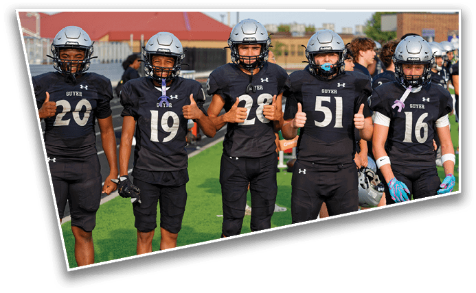 Five people in football uniforms and helmets stand on a field giving thumbs up, with others and buildings visible in the background. Description generated by AI