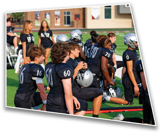 A group of people wearing black football uniforms with helmets are gathered on a green field, some standing and some sitting, with a few people in casual clothing in the background. Description generated by AI