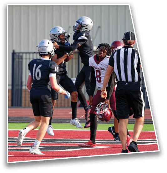 People in football uniforms are on a field with one person holding a helmet and others celebrating or interacting near the end zone. Description generated by AI